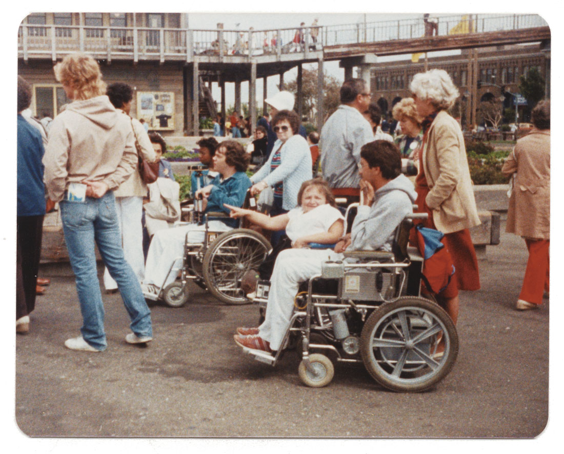 Fotografía a color de un grupo de personas, algunas de pie y algunas en silla de ruedas, en una protesta en San Francisco.
