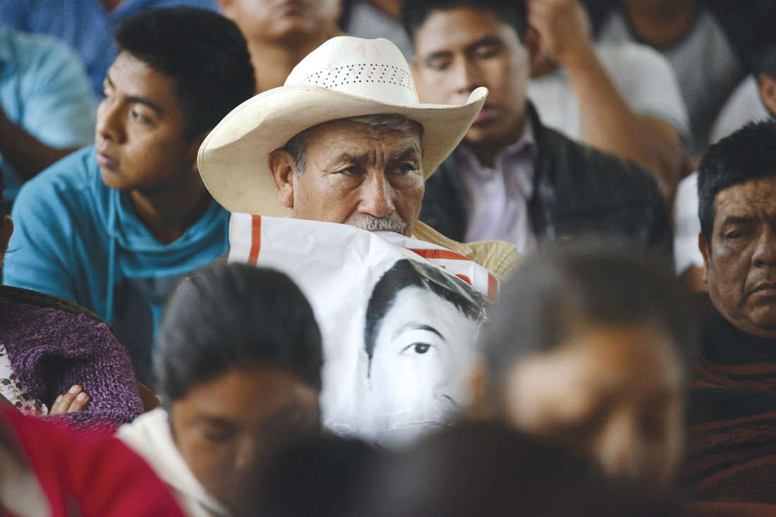 Fotografía de un hombre mayor con un sombrero en el que está escrito el número "43", sostiene la mirada hacia el lado derecho de la imagen, debajo se ve el detalle de una manta impresa con la fotografía de uno de los 43 normalistas desaparecidos de Ayotzinapa.