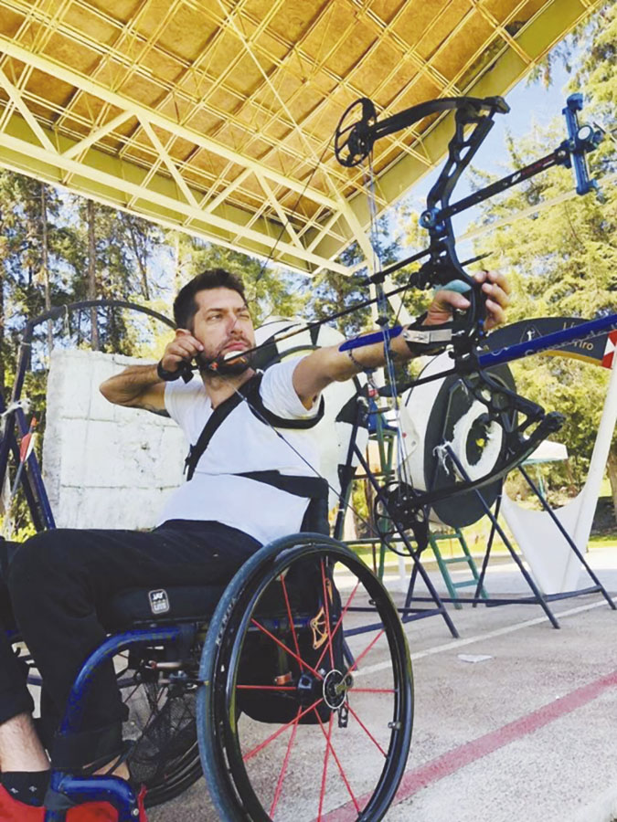 Fotografía de un hombre en silla de ruedas que practica tiro con arco. En la imagen tensa el arco y apunta la flecha en un campo de tiro.