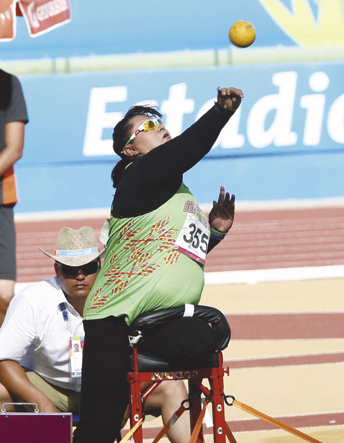 Fotografía de una atleta paralímpica sentada en una plataforma vestida de uniforme verde segundos después de haber arrojado con el brazo extendido una bala que se capta en movimiento en la parte superior derecha de la imagen.