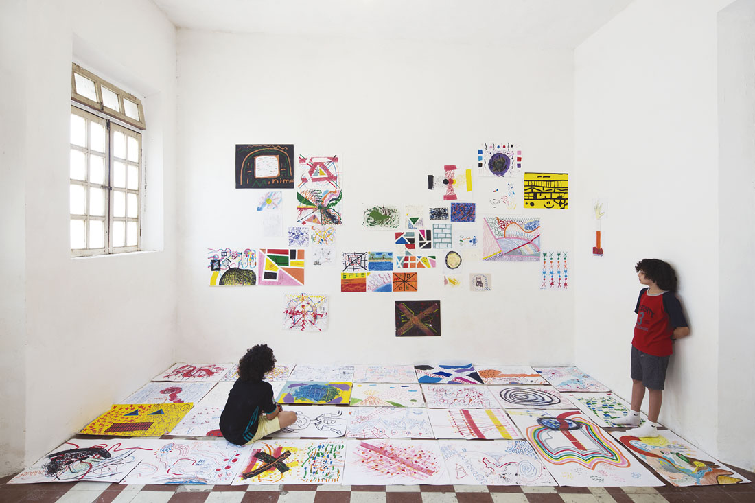 Vista de una exposición con dibujos en hojas colocadas sobre el piso y en el muro. Dos niños de cada lado los observan, uno está sentado y otro de pie.
