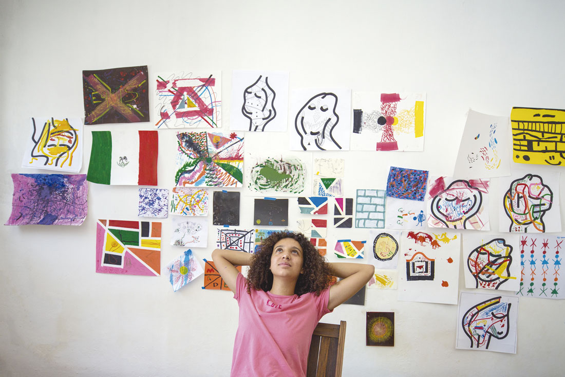 Fotografía de una persona sentada con los brazos detrás de la cabeza y ubicada en frente de una serie de dibujos pegados a la pared.