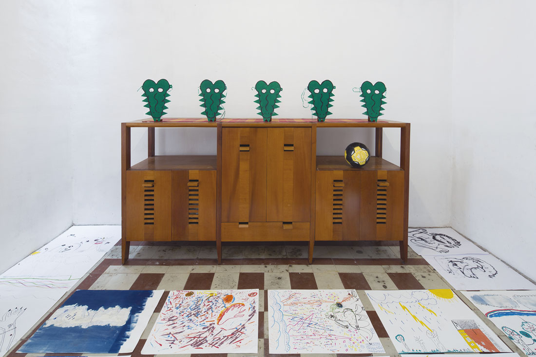Fotografía de una exposición, al fondo un mueble de madera con máscaras de cocodrilo encima y varios dibujos desplegados en el piso.