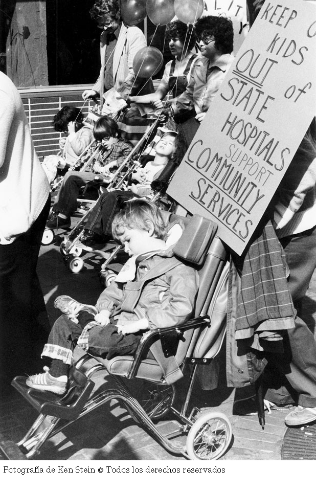 Fotografía en blanco y negro de un grupo de personas en silla de ruedas en una protesta. En el primer plano un niño en silla de ruedas sostiene un cartel que dice "Keep our kids out of state hospitals support community services".