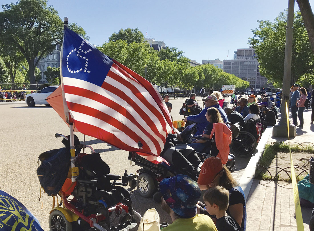 Fotografía de una protesta de personas en silla de ruedas, al frente una bandera de los Estados Unidos que tiene en el cuadrante superior izquierdo una silueta de una persona en silla de ruedas construida  con estrellas blancas sobre fondo azul.