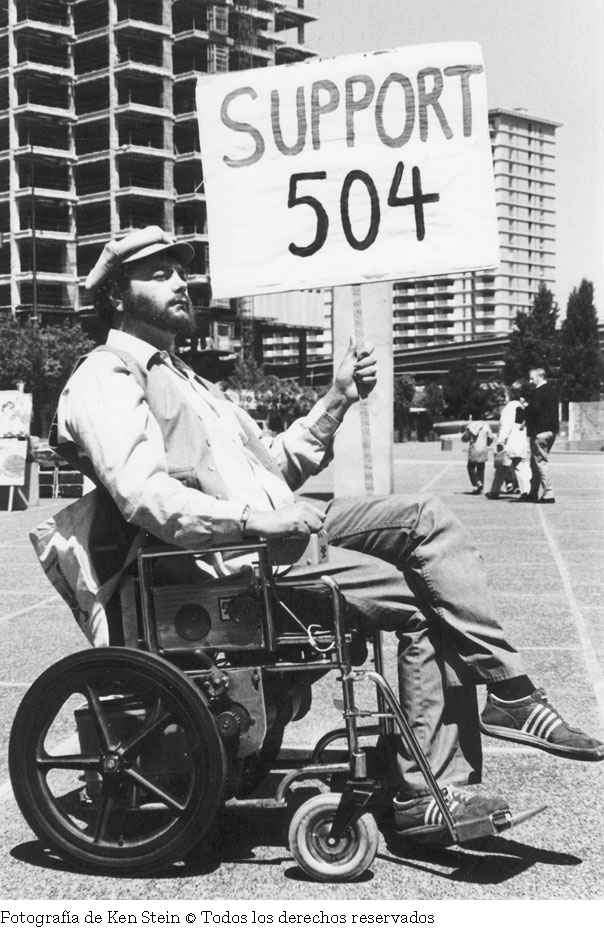 Fotografía en banco y negro de un hombre en silla de ruedas con las piernas cruzadas, sostiene un cartel que dice "Support 504", al fondo edificios en construcción.